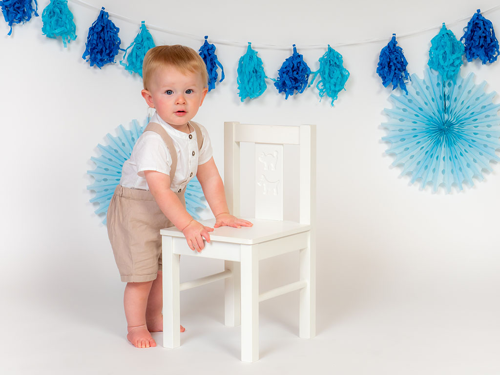 baby boy standing by white chair before cake smash taken by baby photographer in Braintree